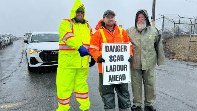 Three workers standing outside on a road blocking a vehicle holding a sign that reads warning scab labour ahead.